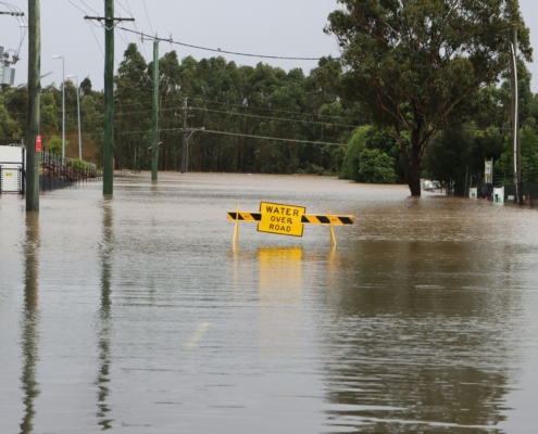 A flooded road lined with utility poles and trees. In the center, a yellow emergency barricade with the text "WATER OVER ROAD" stands in murky, brown water that has completely covered the asphalt.