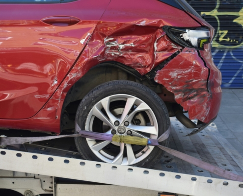 Side view of a red hatchback on a tow truck showing severe collision damage. The metal above the rear wheel is crumpled, the taillight is smashed, and the rear bumper is partially detached. Purple heavy-duty straps secure the rear wheel to the flatbed.