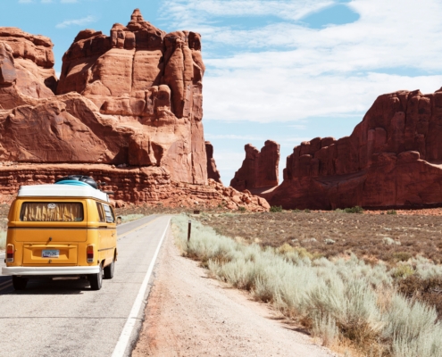Rear view of a vintage yellow camper van driving on an open desert road toward massive red rock formations under a blue sky.