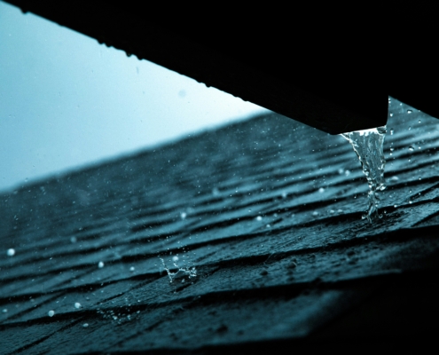 Close-up view of heavy rain pouring off a dark roof gutter onto textured roof shingles during a storm, with water droplets splashing under a moody blue sky.